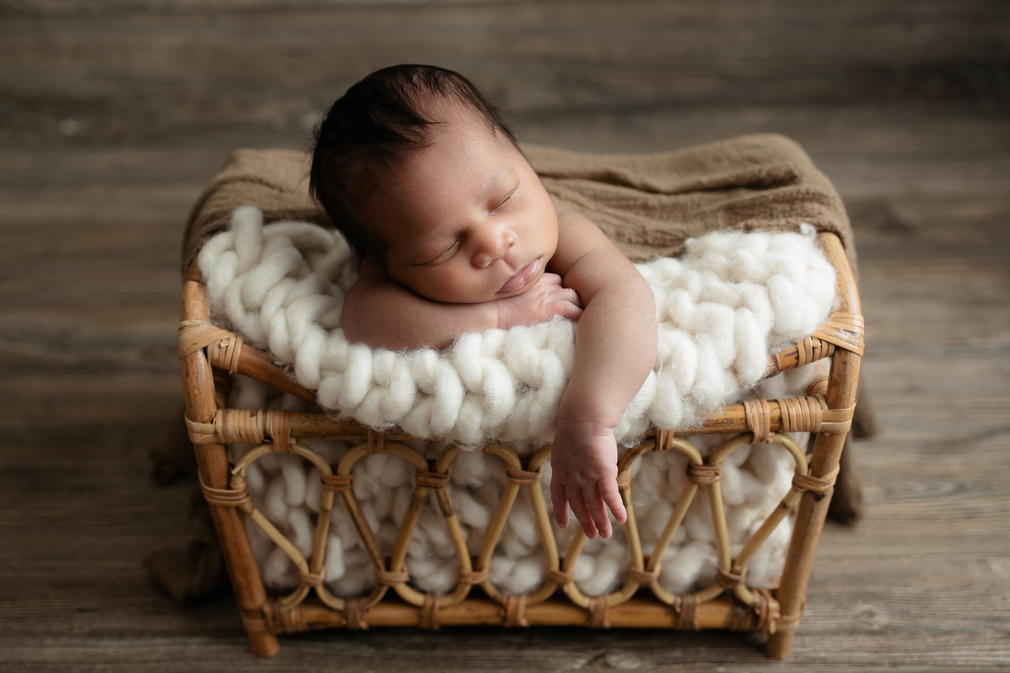 Newborn baby boy sleeping in a neutral taupe wrap, posed safely in a rustic wooden basket at our Manassas, Virginia studio.