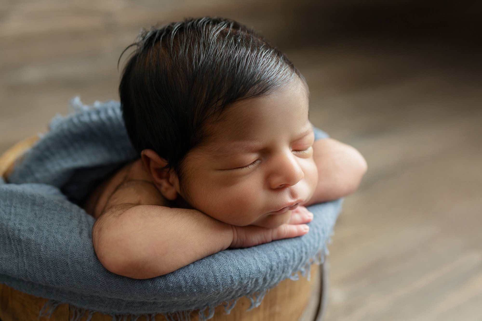 Newborn baby boy sleeping soundly in a neutral taupe wrap, posed safely in a rustic wooden bowl at our Independent Hill studio.