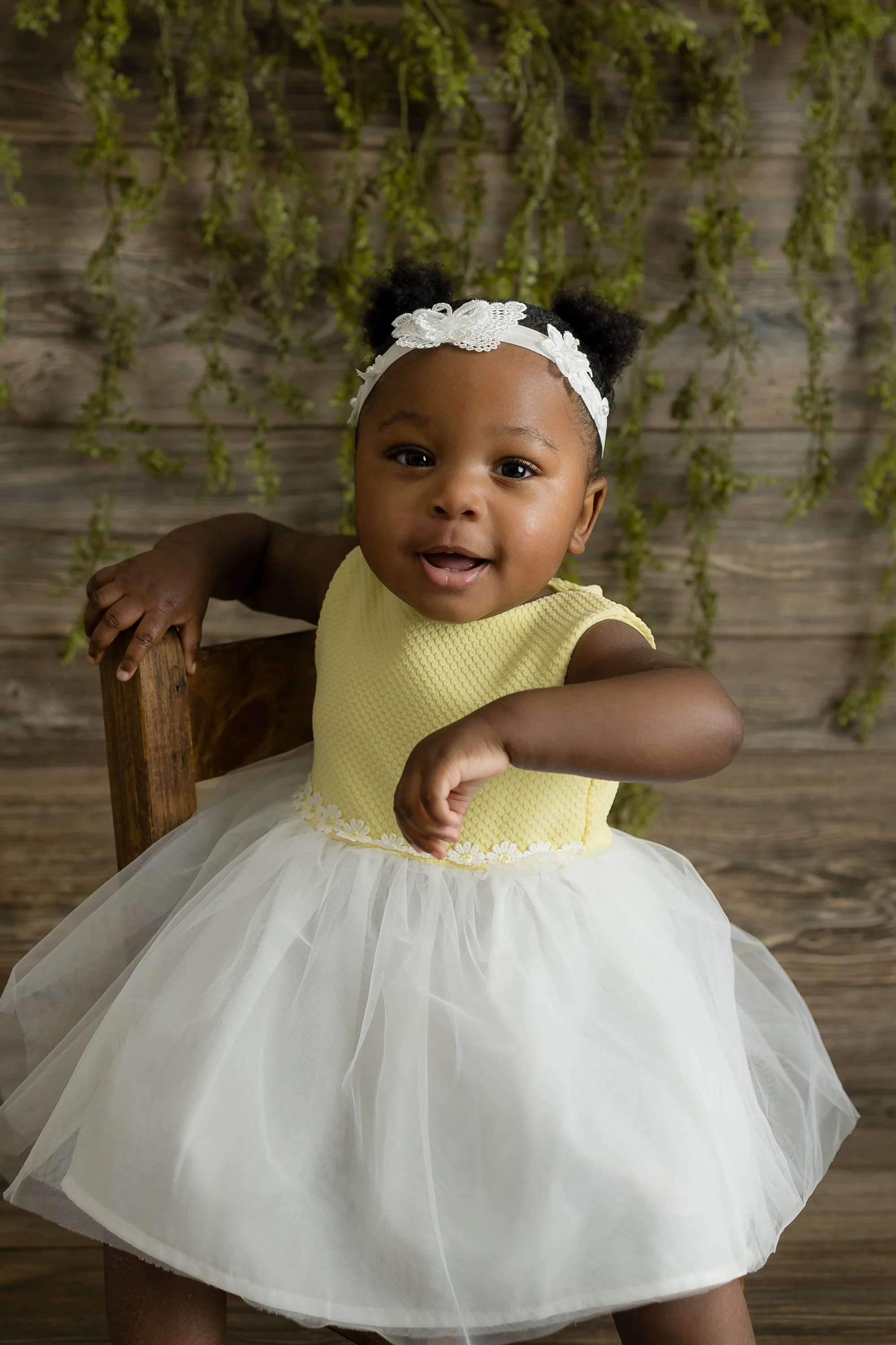 A happy one-year-old baby girl smiling and sitting in a bright studio setup, celebrating her first birthday milestone with a small cake and floral decorations in Northern Virginia.
