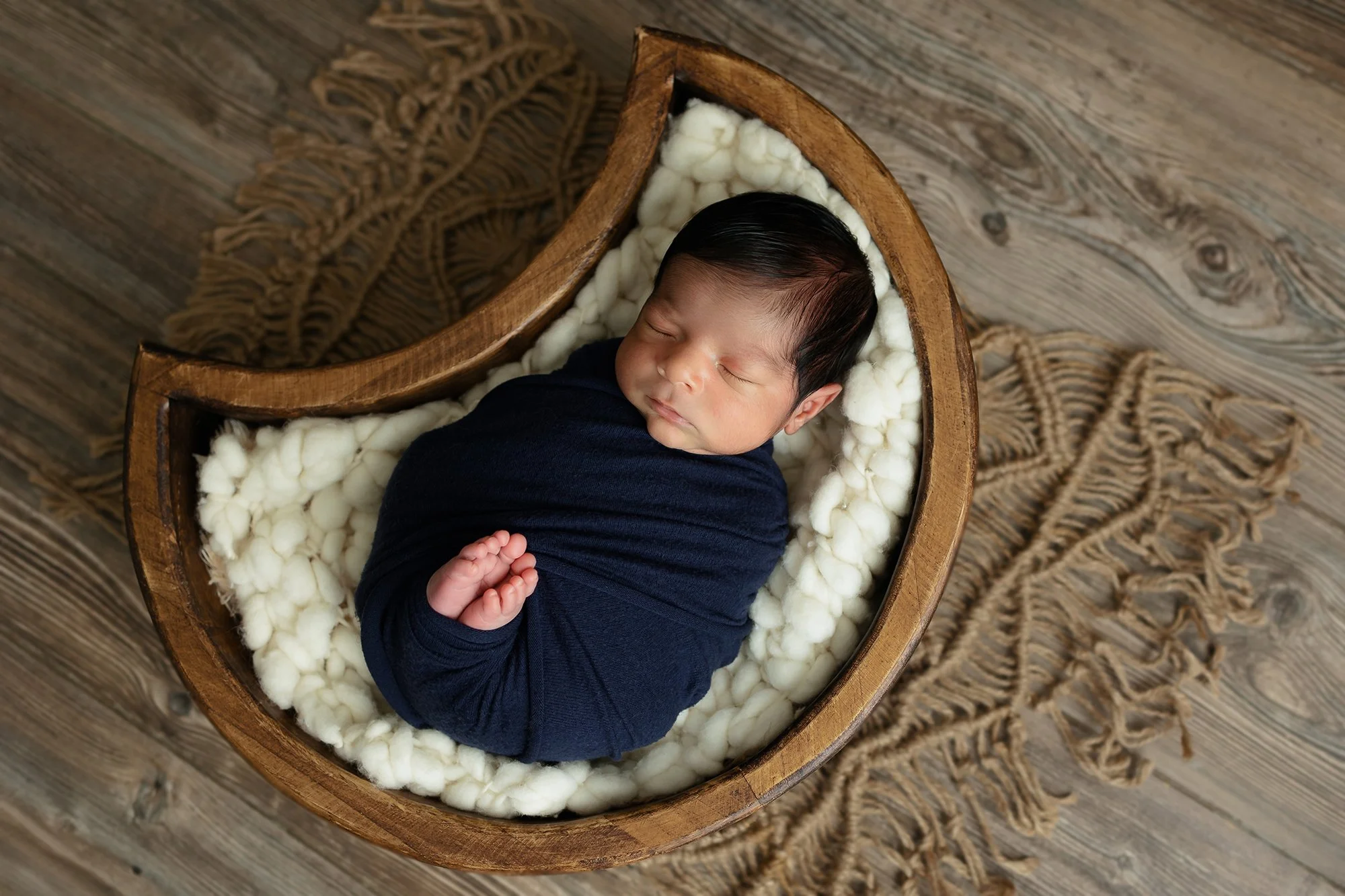 Newborn baby safely curled in a vintage-style wooden heart bowl with organic textures, a signature petite session look.