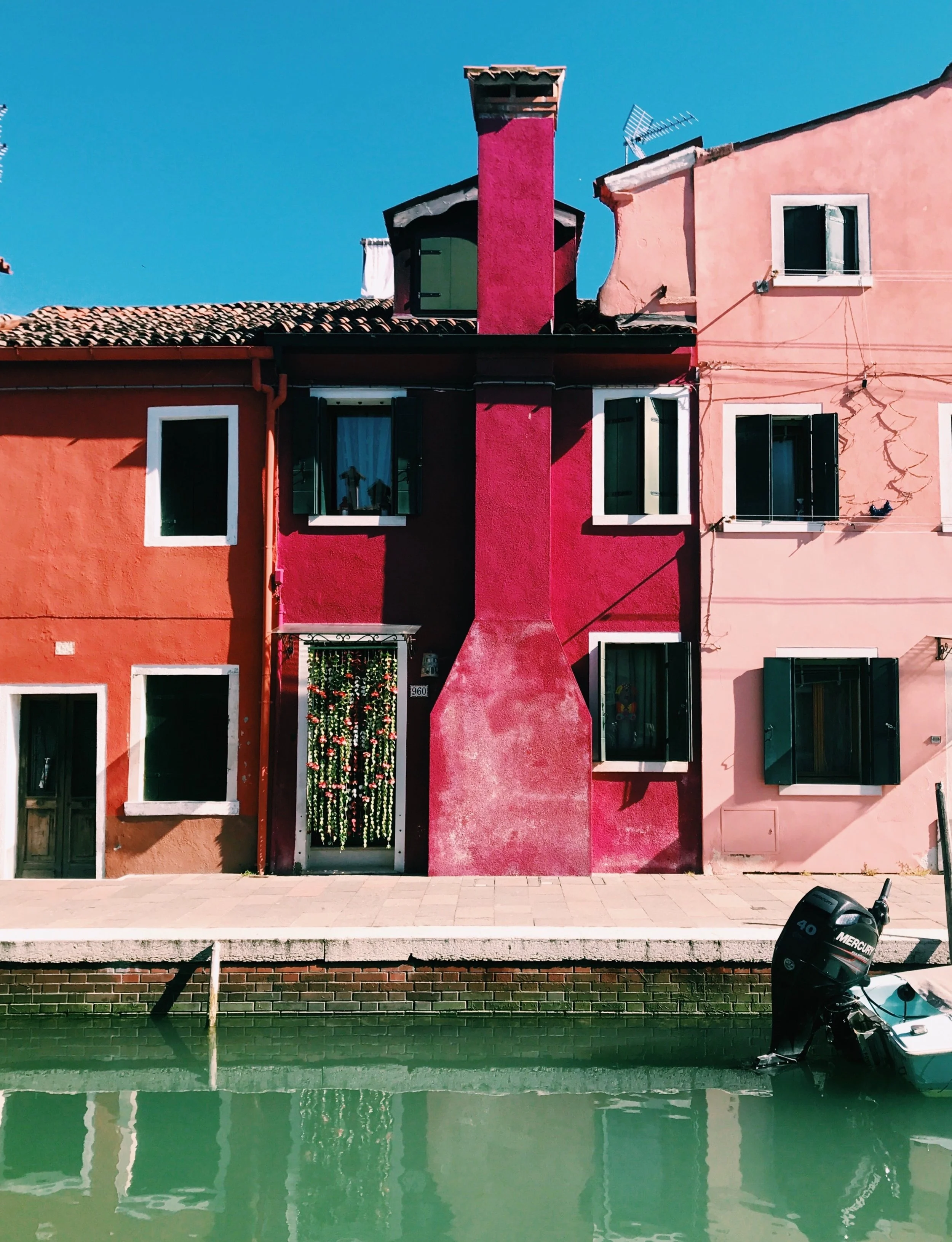Colorful houses with a canal in front, featuring vibrant pink and orange walls, white window frames, and a small boat with an outboard motor on the water. The sky is clear and blue.