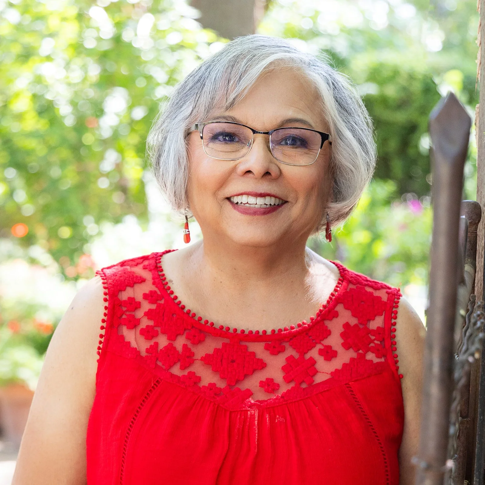 Woman with glasses smiling, wearing a red sleeveless top with floral embroidery, standing outdoors with greenery in the background.