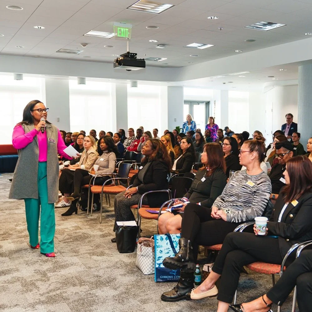 A woman in a pink blazer and teal pants speaking into a microphone during a conference or seminar with a seated audience of diverse women and a few men in a bright, modern room.