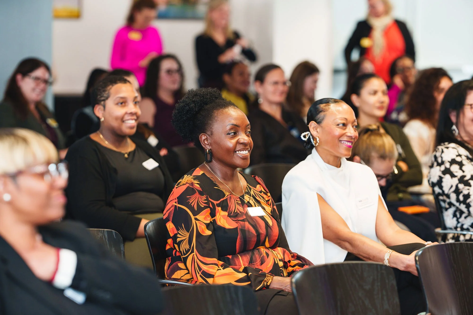 Group of diverse women seated and smiling at a conference or seminar, with some women standing in the background.