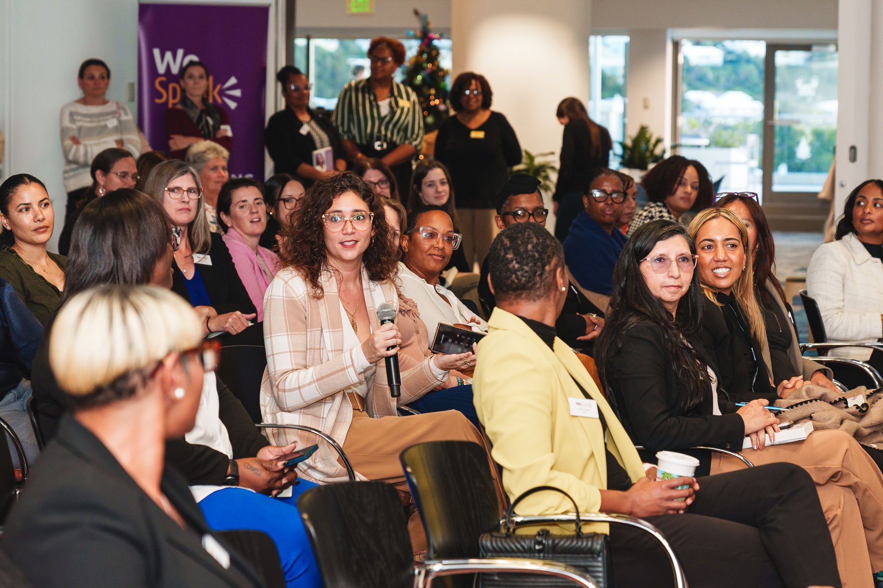A woman in a plaid blazer sits among a diverse group of people in an audience, holding a microphone and a tablet, in a conference room decorated with a Christmas tree and a purple banner that reads 'We Speak.'