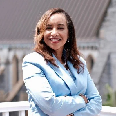 A woman in a light blue blazer smiling with arms crossed outdoors.