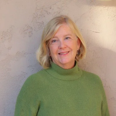 A smiling older woman with blonde hair wearing a green turtleneck sweater, standing against a textured light-colored wall.