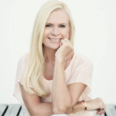 A smiling woman with long blonde hair, wearing a light-colored top, resting her chin on her hand, in front of a white background.