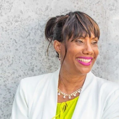 A woman with brown hair tied in a ponytail, wearing a white blazer and a bright yellow top, smiling and looking to the side against a textured gray background.