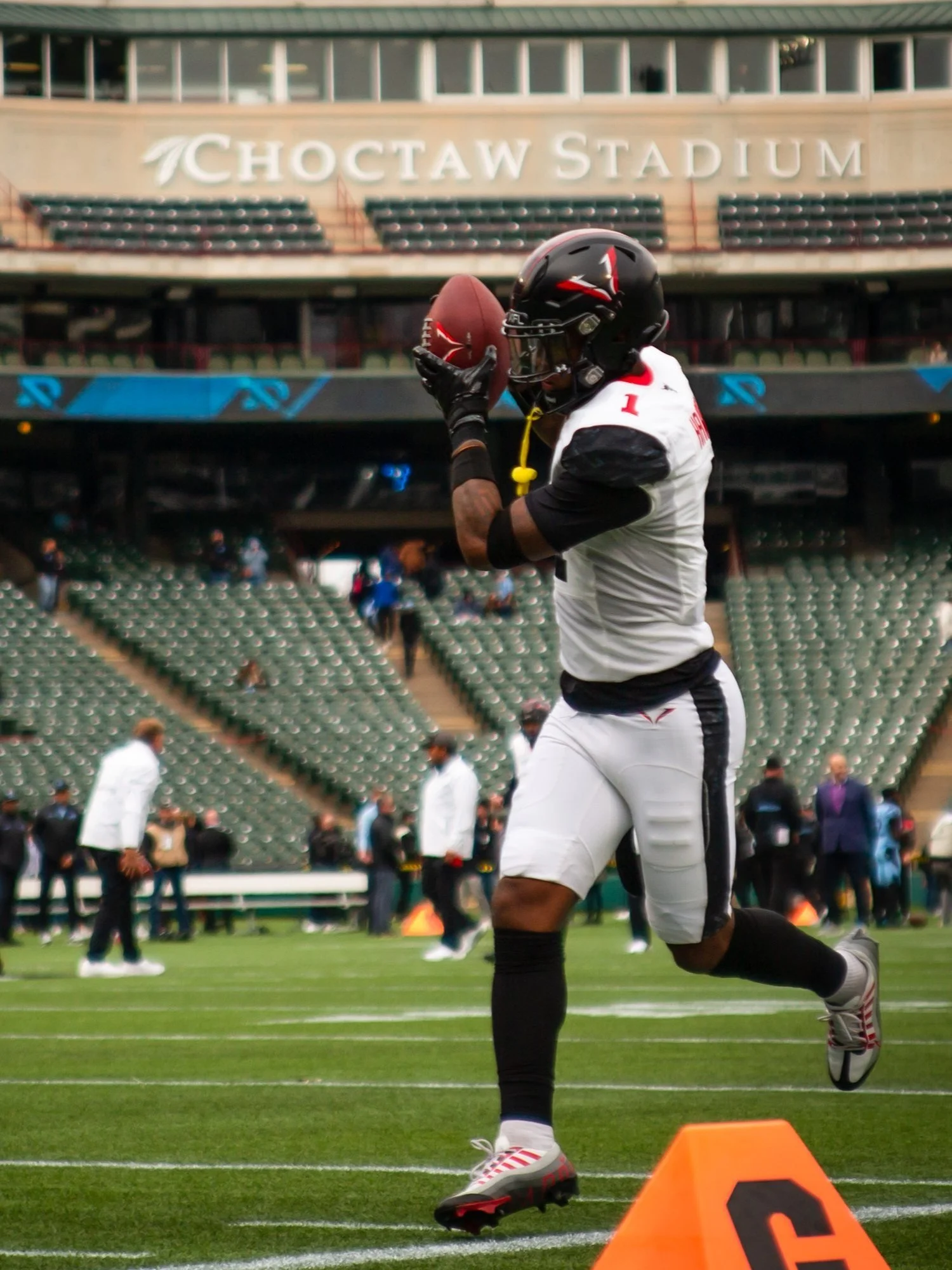 An American football player wearing a black helmet, white jersey, and black and white pants holding a football in a stadium.
