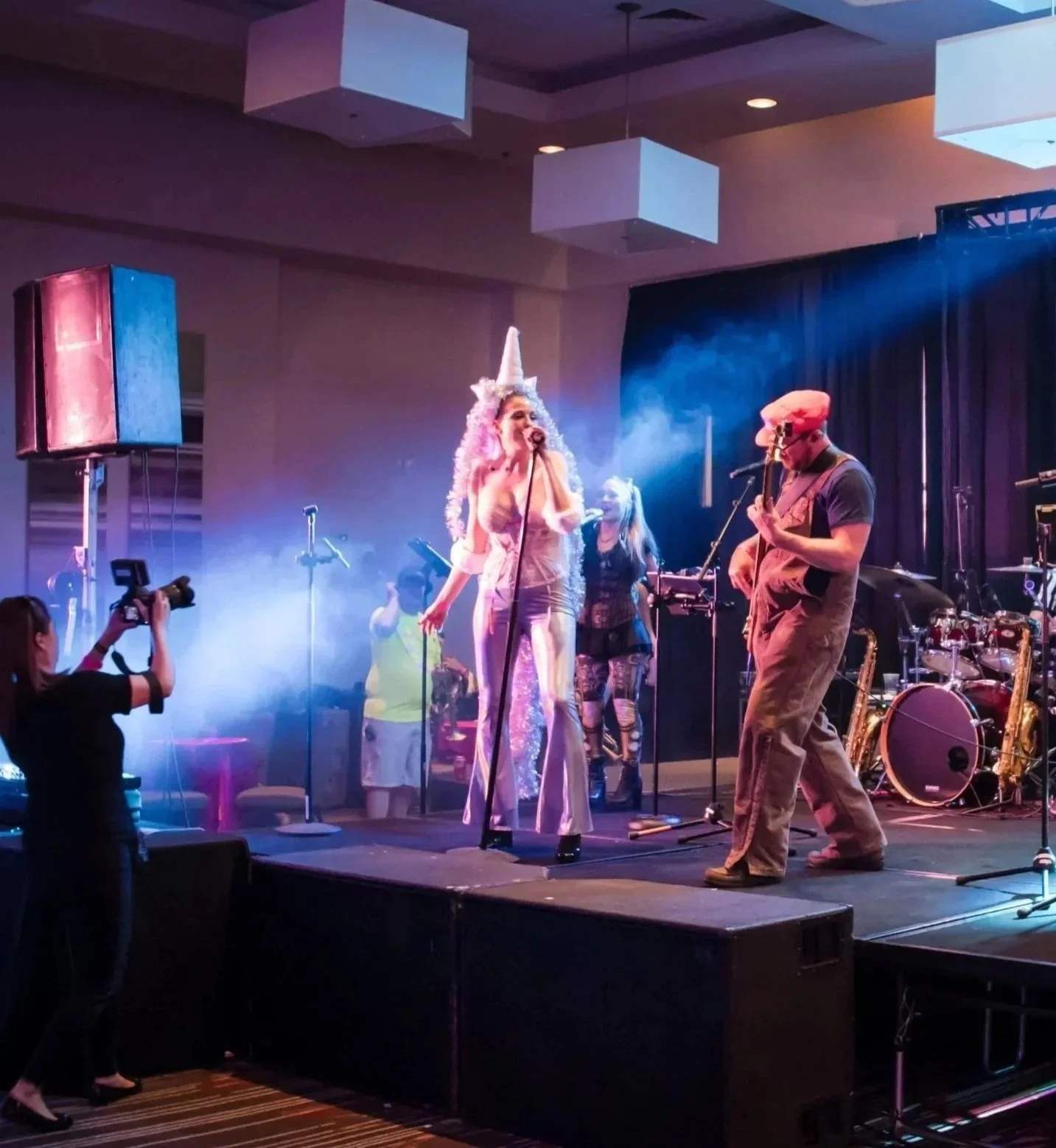 A live music performance on stage with various musicians, including a singer in a unicorn costume, a guitarist wearing a cap, a pianist, and other band members under colorful stage lights. An audience member is taking photos.