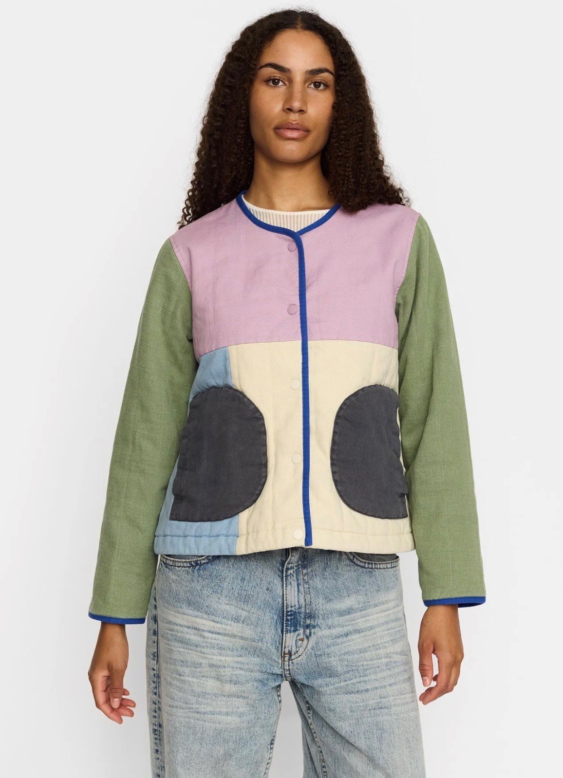 A woman wearing a colorful patchwork quilted jacket with various pastel and dark colors and light blue jeans.