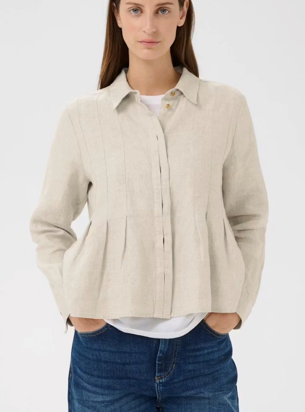 Woman wearing a light beige blazer over a white shirt, with hands in her pockets, standing against a plain background.