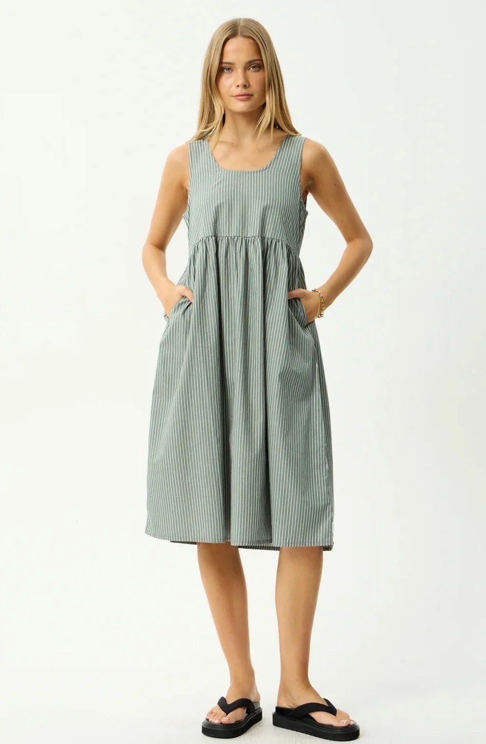 A woman wearing a sleeveless gray and white striped midi dress with pockets, standing against a plain white background.