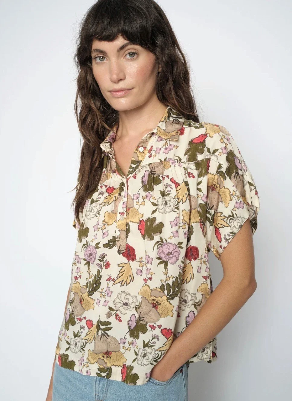 A woman with long wavy dark hair and fair skin, wearing a floral patterned beige blouse with short sleeves and a collar, standing against a plain white background.