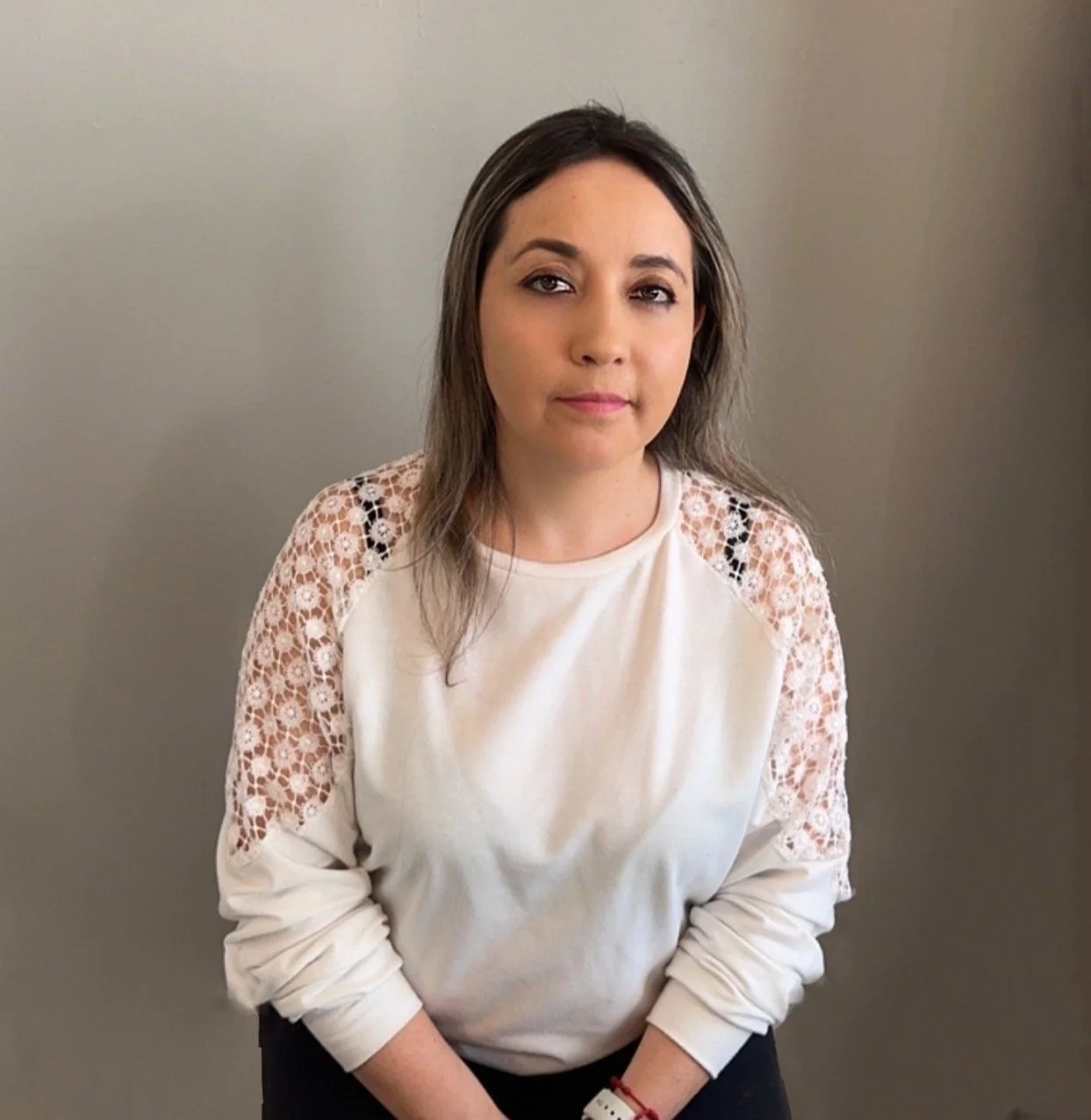 A young woman, named Lorena, with long brown hair and light skin, wearing a white long-sleeve shirt with lace detailing on the shoulders, standing against a plain gray wall. Founder of Sweet Dream Clean.