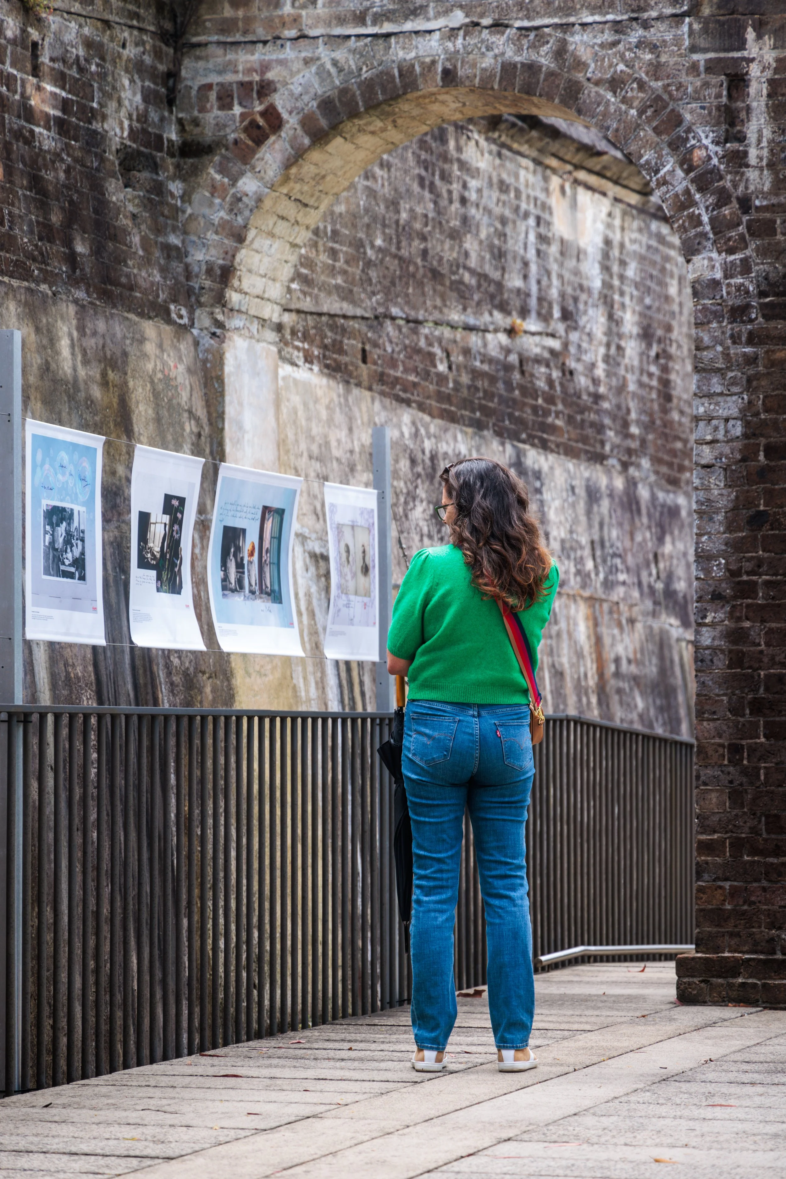 A woman with curly hair and glasses wearing a green sweater and blue jeans, standing outdoors near a brick wall and looking at display panels with pictures and text.