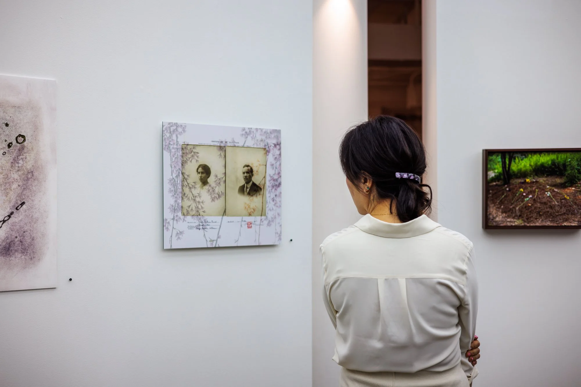 A woman with dark hair styled in a bun and wearing earrings and a cream-colored shirt is standing in front of a white wall, looking at framed artwork displayed on the wall in an art gallery.