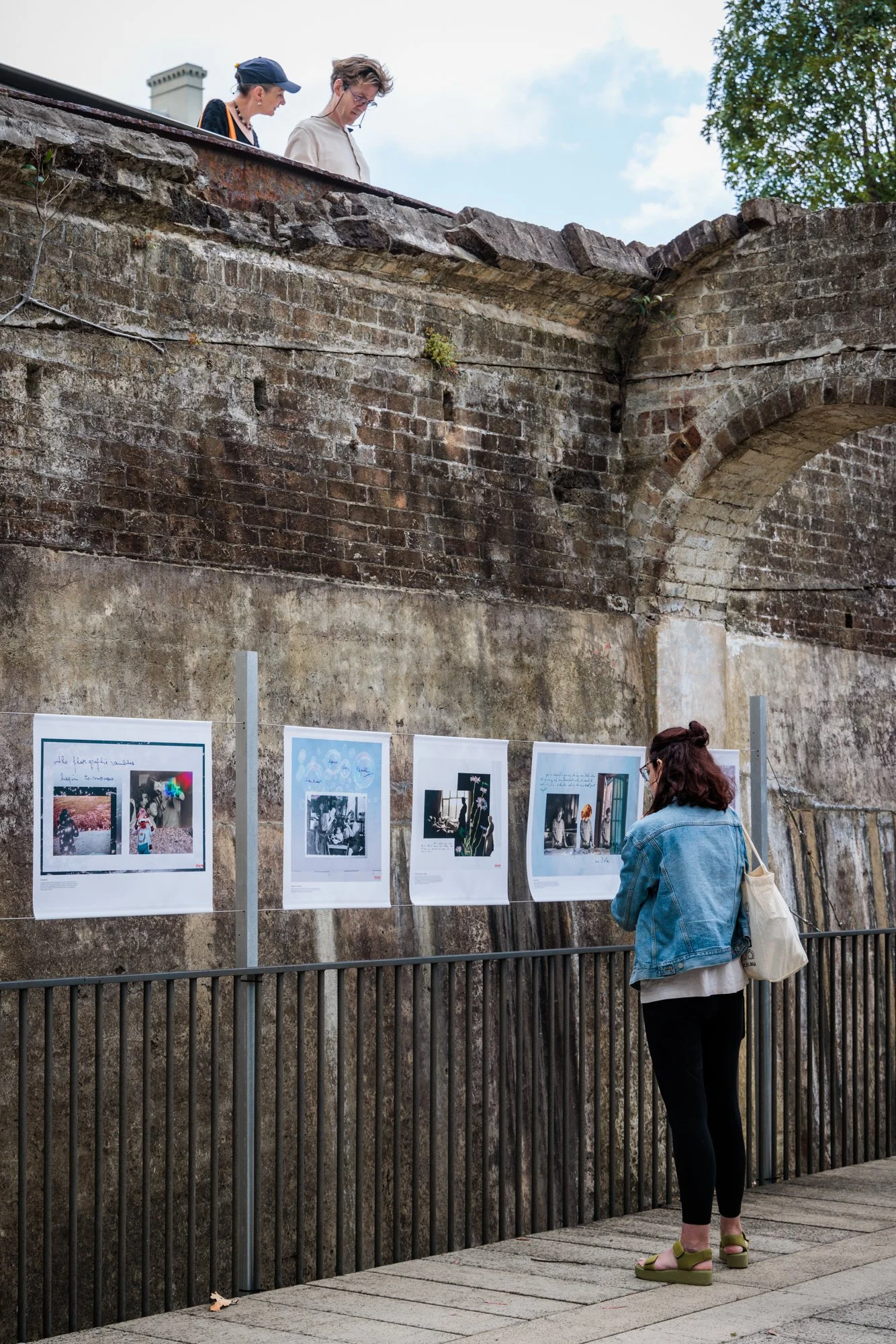 A woman in a denim jacket, black pants, and green platform sandals looks at photographs displayed on a wall outdoors, with rusty brick and stone structures in the background.