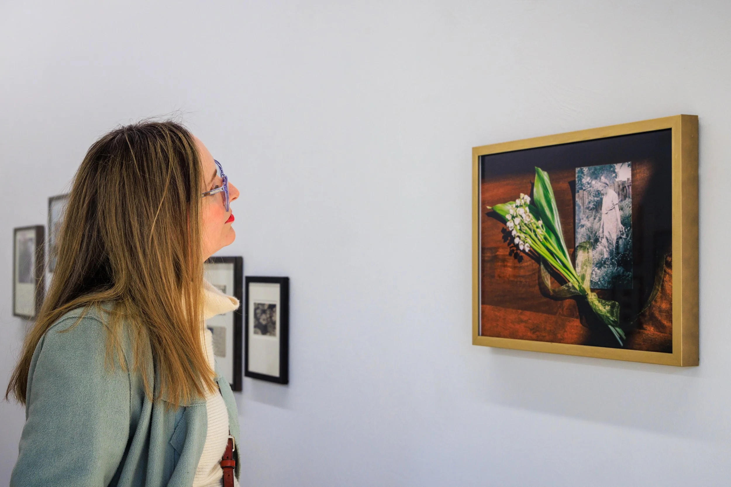 A woman with long hair and glasses looking at a framed artwork of flowers inside an art gallery.