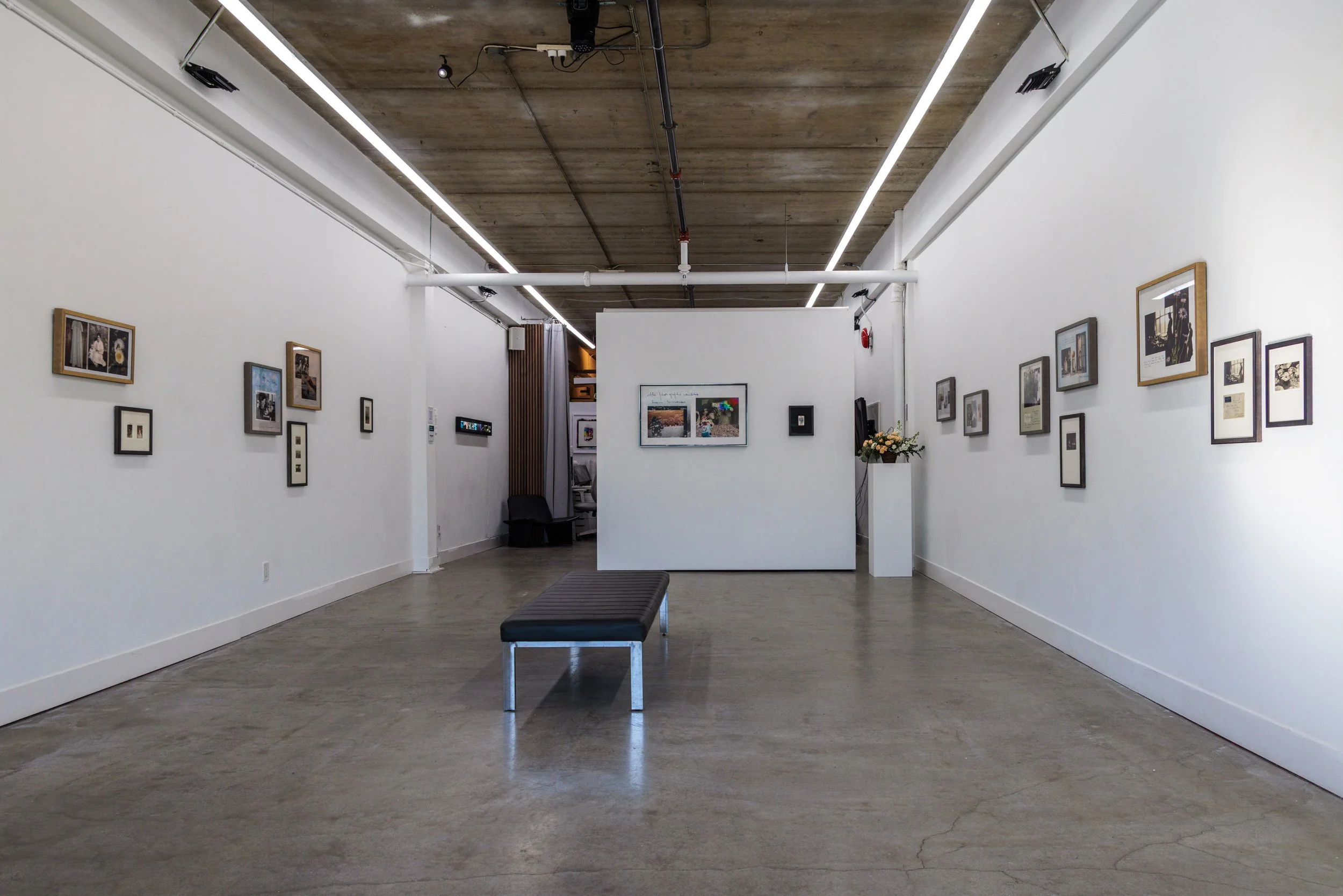 An art gallery with white walls, concrete ceiling, and framed artwork displayed on the walls. A black bench is centered on a polished concrete floor.