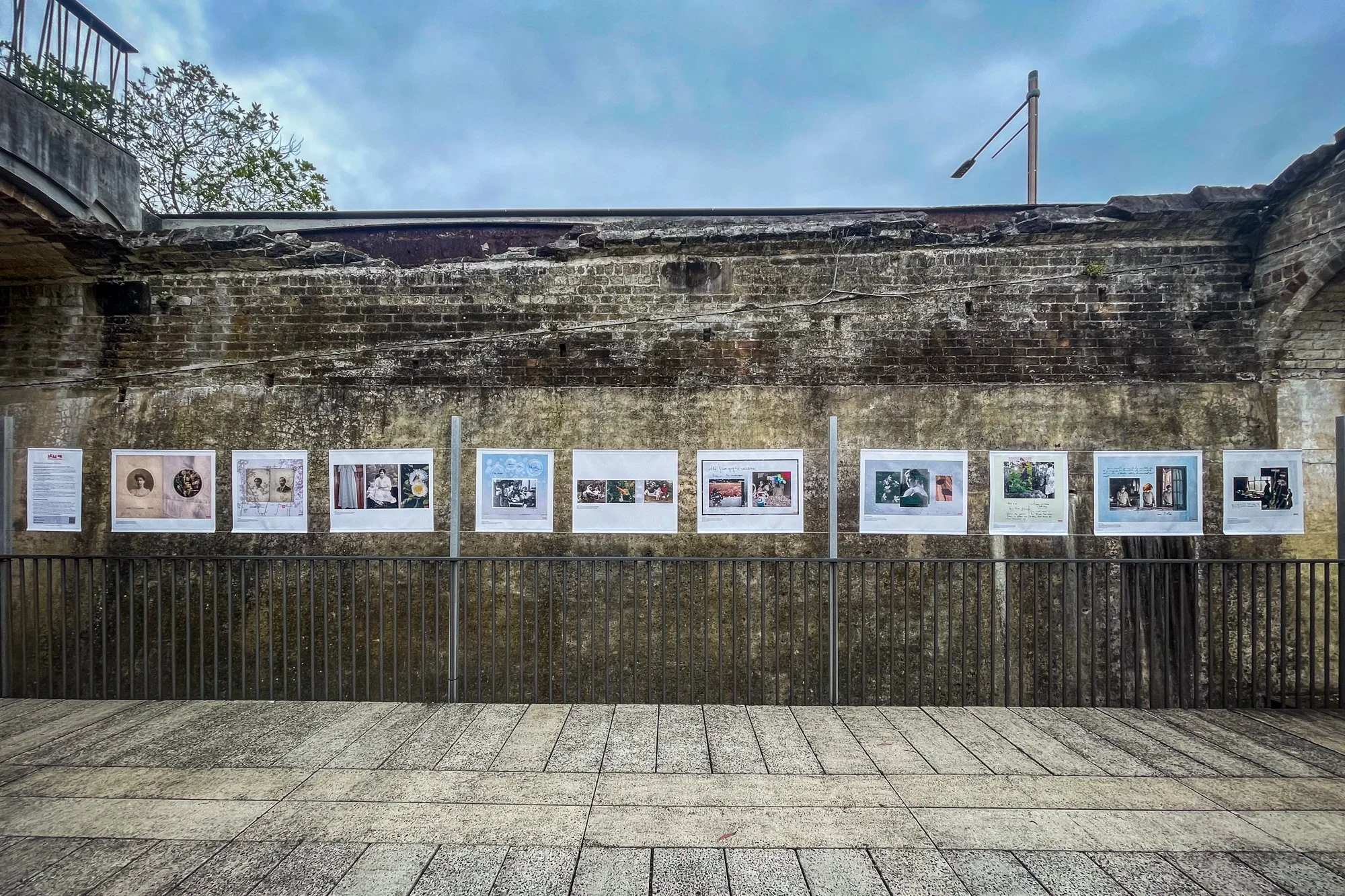 An outdoor display of photographs mounted on a weathered brick wall with an iron railing in front. The wall is part of an old stone structure.