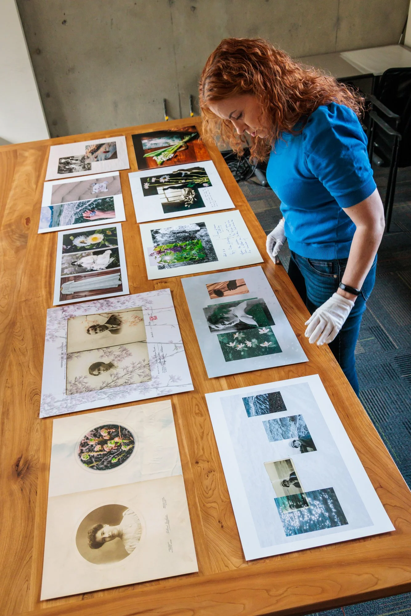 A woman with curly red hair and white gloves is examining various printed photographs and images laid out on a wooden table. The images include portraits, nature scenes, flowers, and artistic compositions.