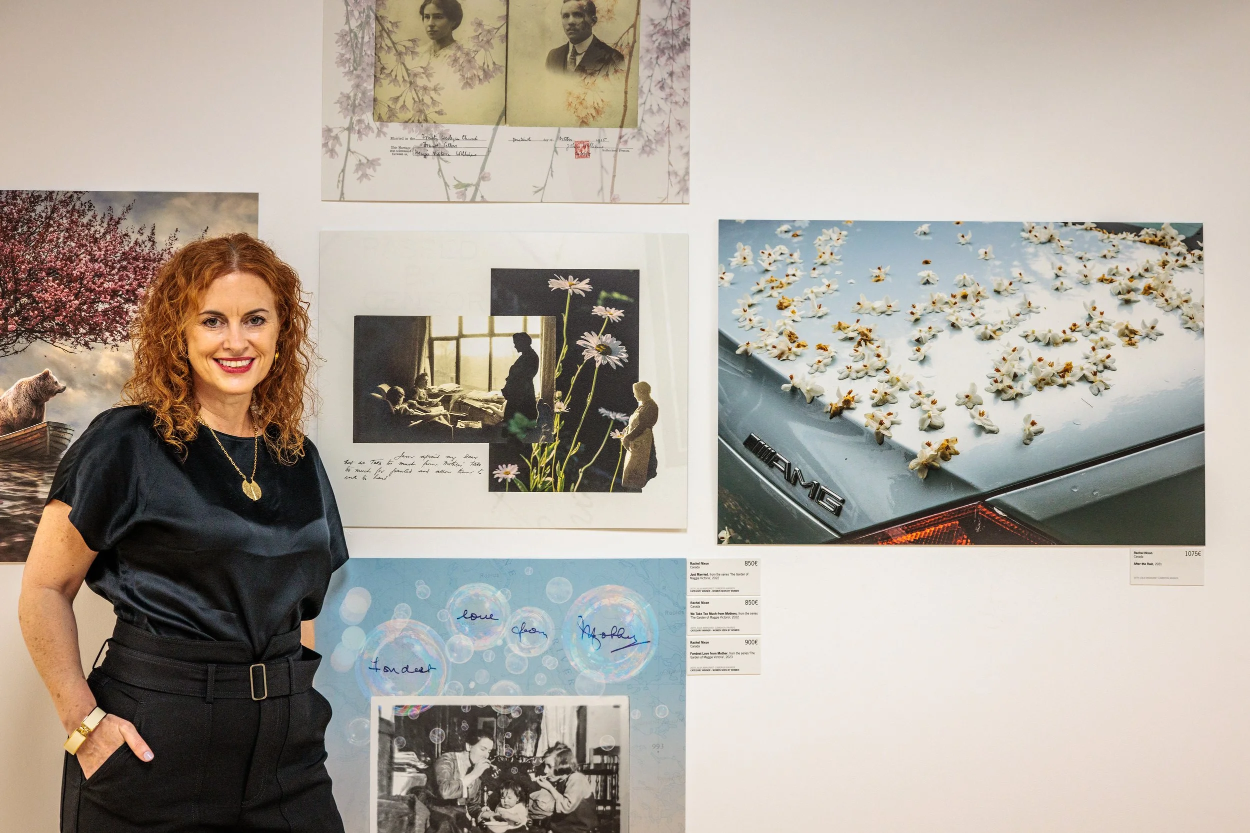 A woman with curly red hair wearing a black top and a gold necklace, smiling in an art gallery with photographs and artwork on the wall behind her.