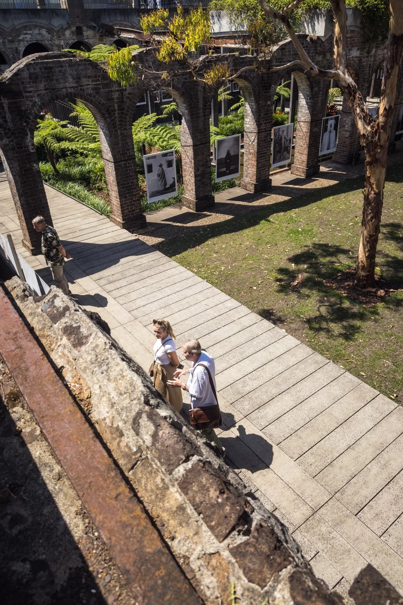 People walking and viewing photographs displayed on a brick wall in an outdoor garden area with greenery and trees.