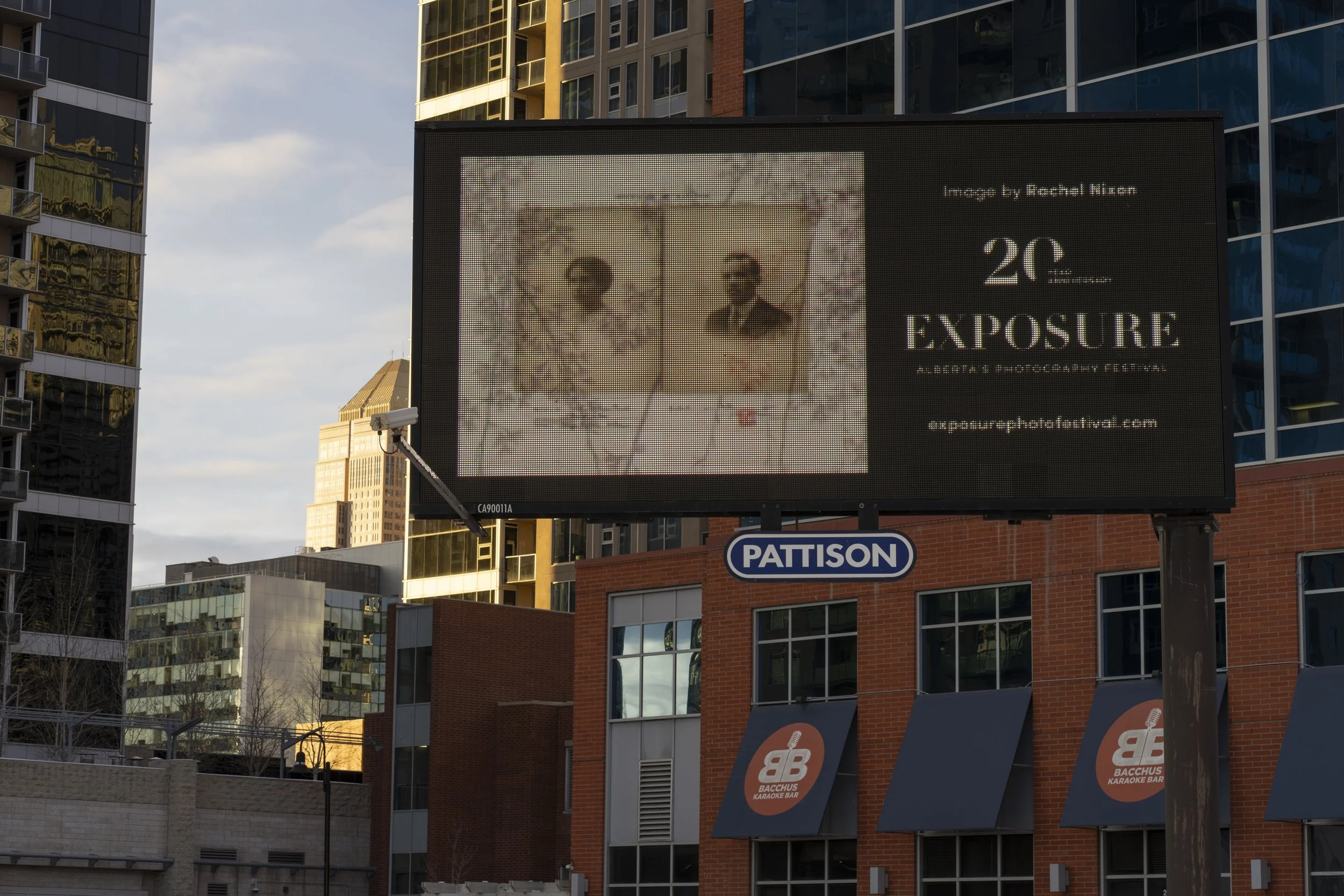 Digital billboard advertising the 20th Exposure Photography Festival, with an image of two people and text including the festival's name and website, set against modern city buildings.