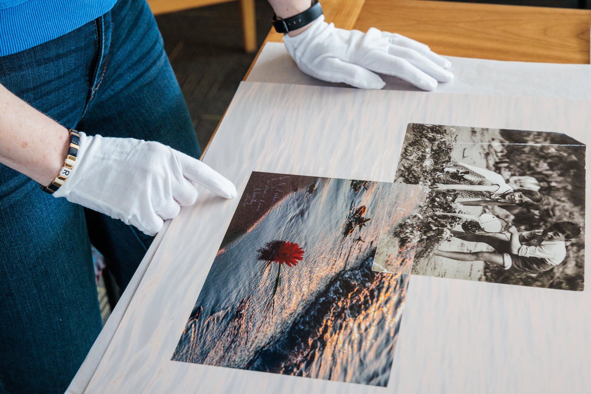 Person wearing white gloves and a black watch looking at printed photographs on a table. The photographs include a sunset over water with a flower and a black-and-white picture of children at the beach.