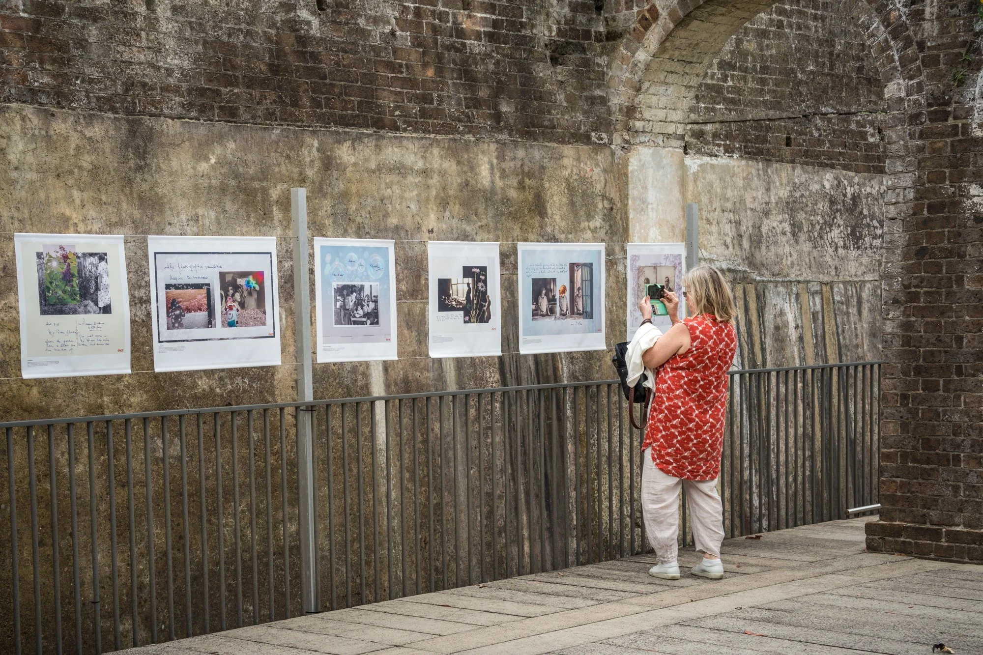 A woman taking a photo of an art display on a brick wall in an outdoor exhibition. She is wearing a red and white patterned sleeveless shirt and white pants.