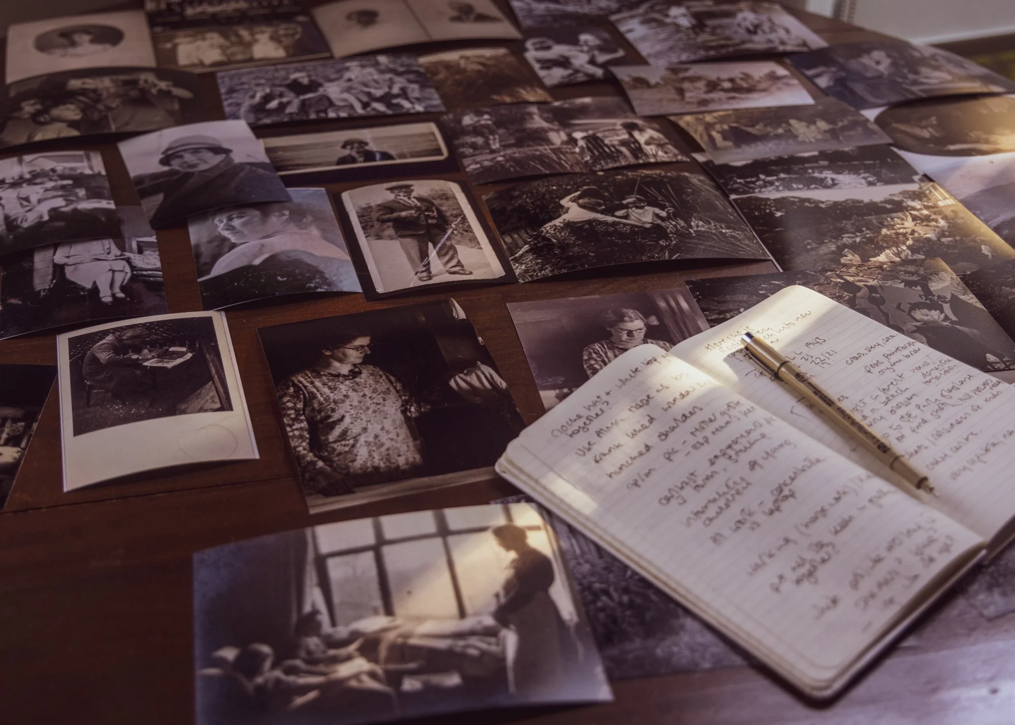 A collection of black and white photographs spread out on a table, alongside a handwritten notebook with a pen resting on it. The photos appear to be vintage images of people and scenes, and the notebook has cursive writing.