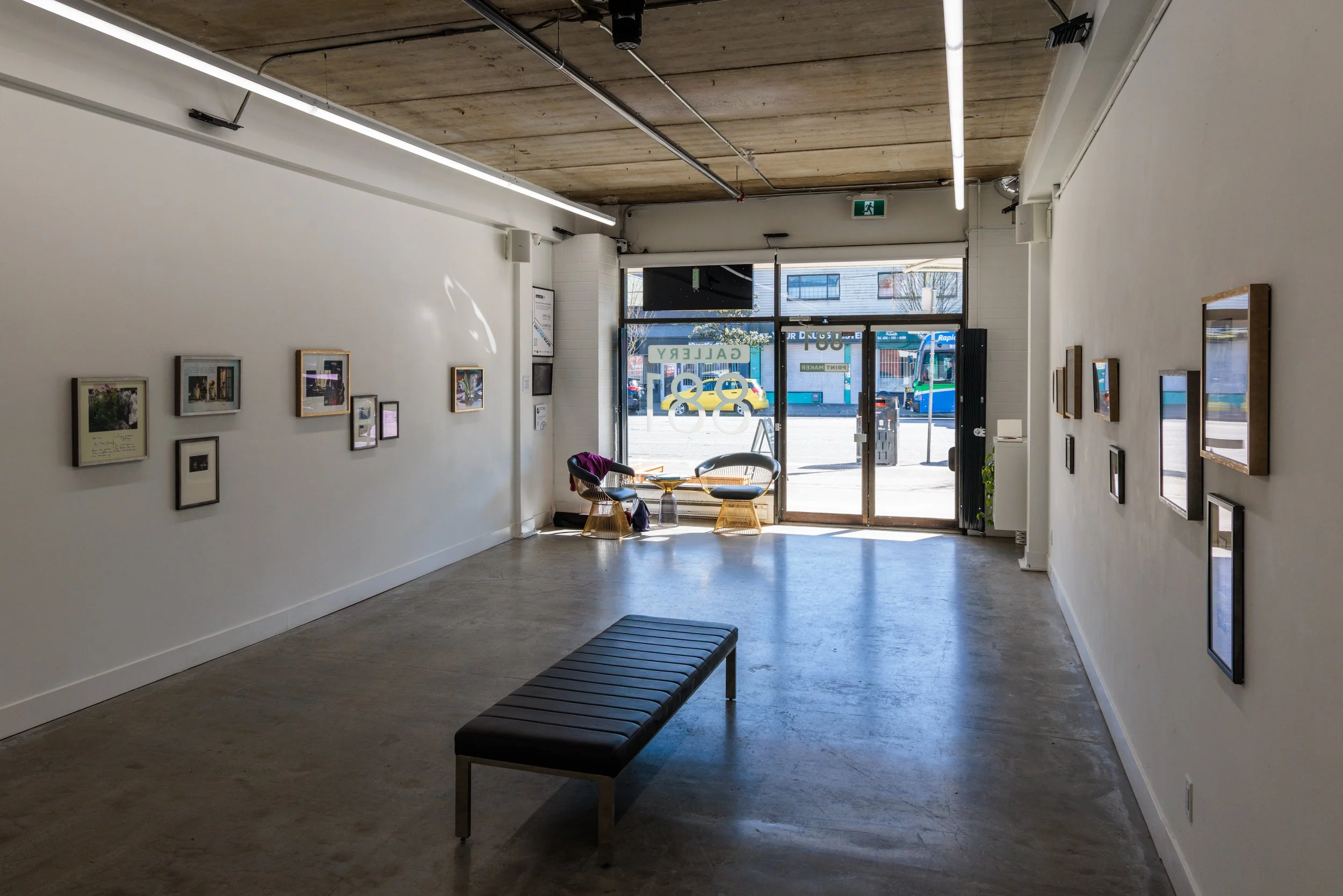 A gallery with framed pictures on white walls and a black bench in the center. There are chairs near the entrance, which has glass doors showing city street outside.