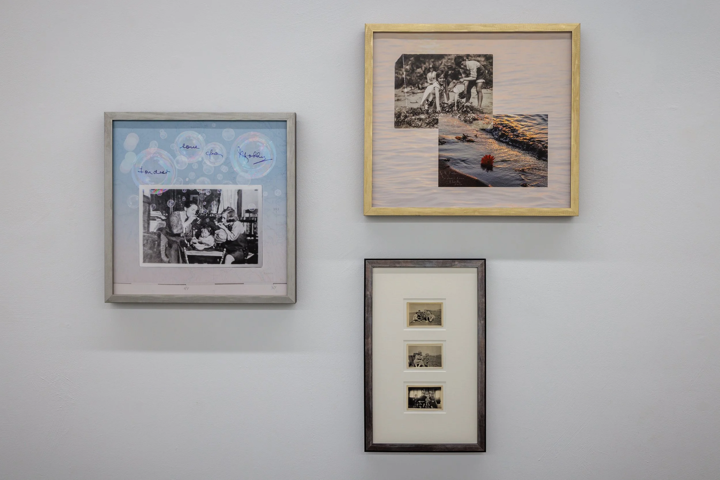 A wall with several framed photographs and artwork. The largest frame contains a collage of black-and-white images of children playing by the water and on a playground, with a background of water at sunset. Adjacent frames show a vintage photo of a w