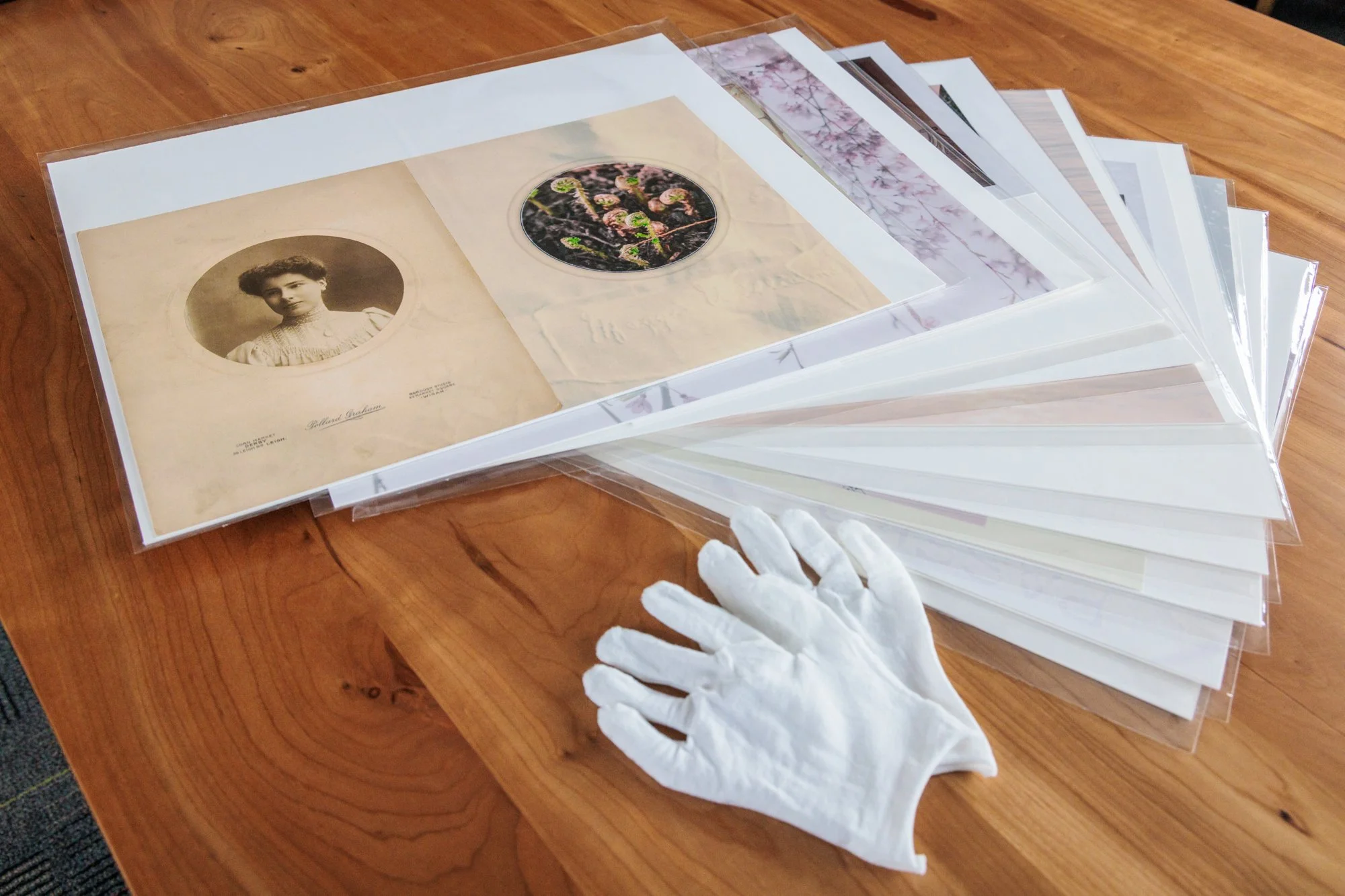 A stack of plastic sleeves holding large printed photo collages, with a pair of white gloves resting on the table.