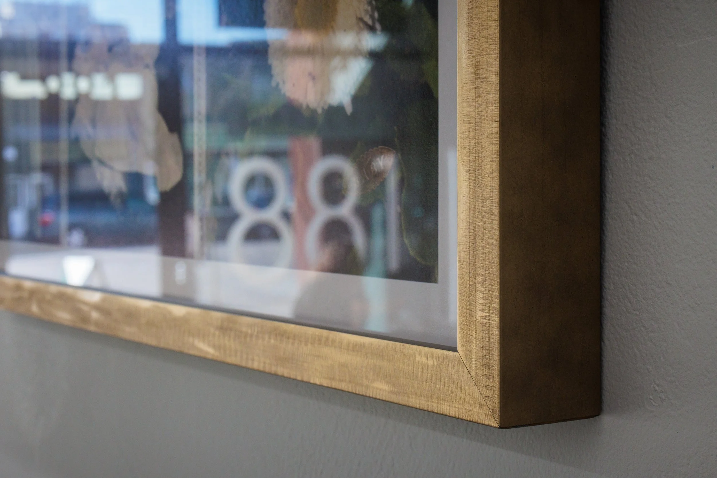 A framed picture hangs on a wall, with the frame made of wood and the picture featuring abstract or blurred images, possibly of a landscape or cityscape, with a muted color palette.