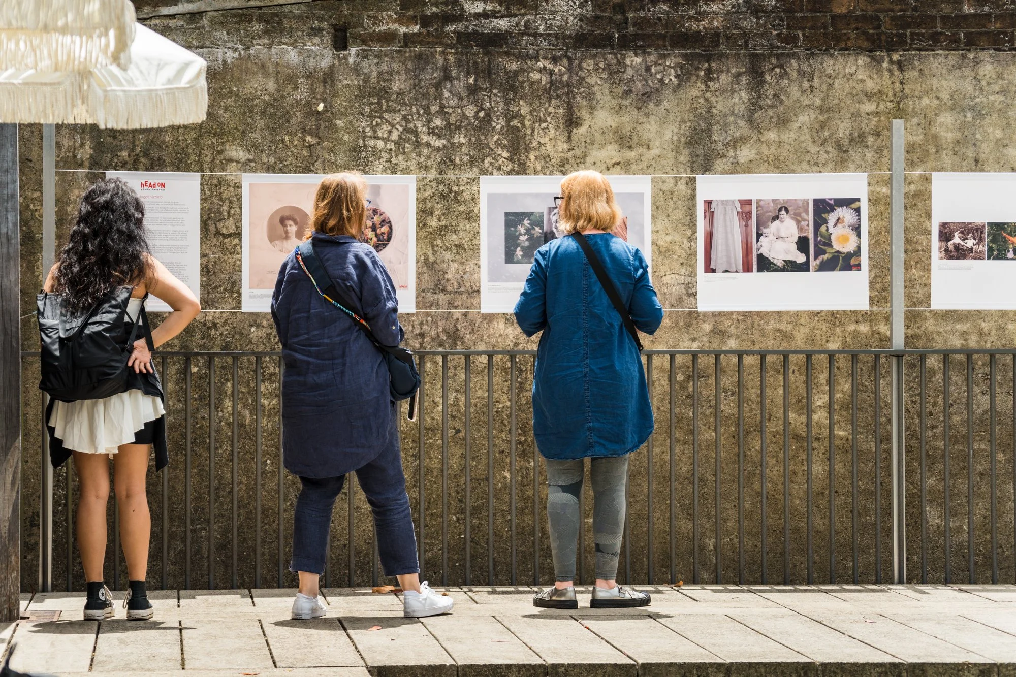 Three women standing in front of a photo display on a brick wall, viewing pictures at an outdoor art exhibit. 