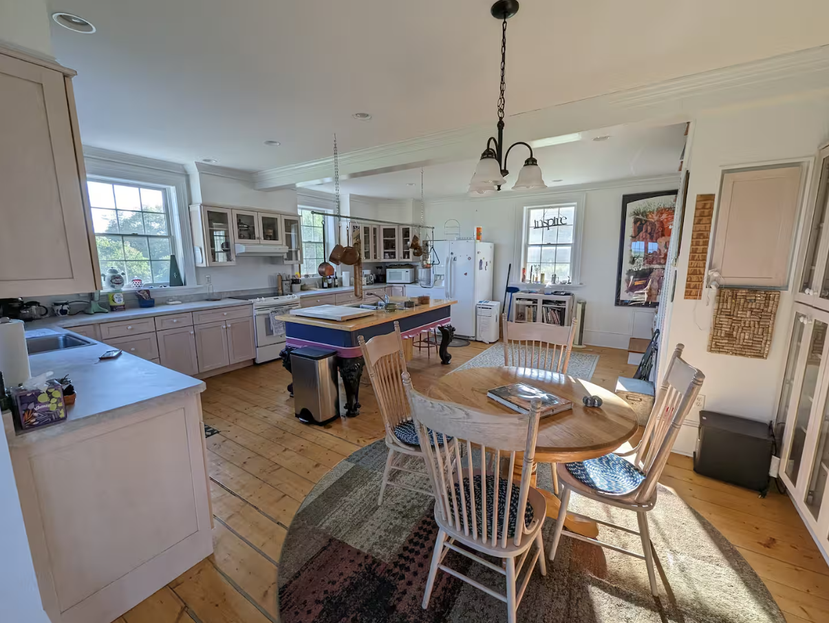 Bright kitchen with white cabinets, a wooden dining table with chairs, and a central island. Windows let in natural light, and various kitchen appliances and utensils are visible.
