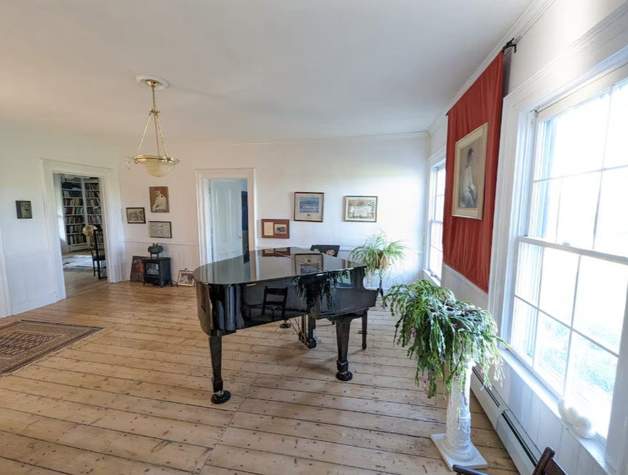 A spacious room with a black grand piano in the center, large windows with sheer curtains, decorated with framed paintings and plants, and a doorway leading to another room with a bookshelf.