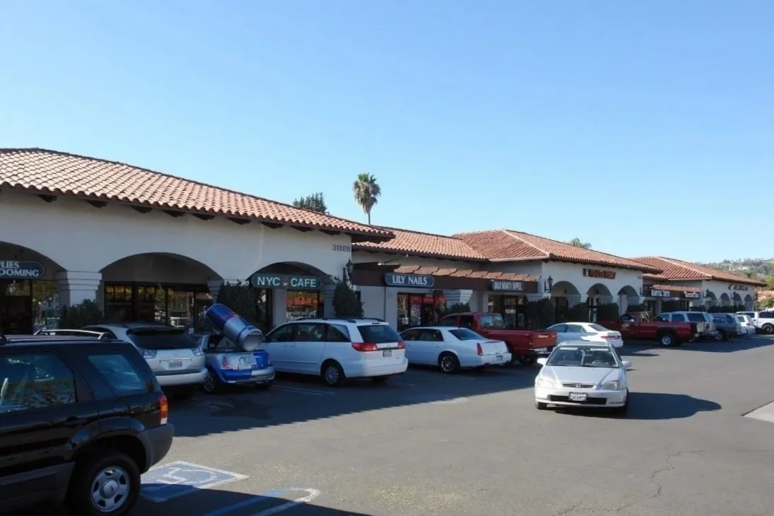 Strip mall with various storefronts including a cafe, nail salon, and other shops, with parked cars and a clear blue sky.