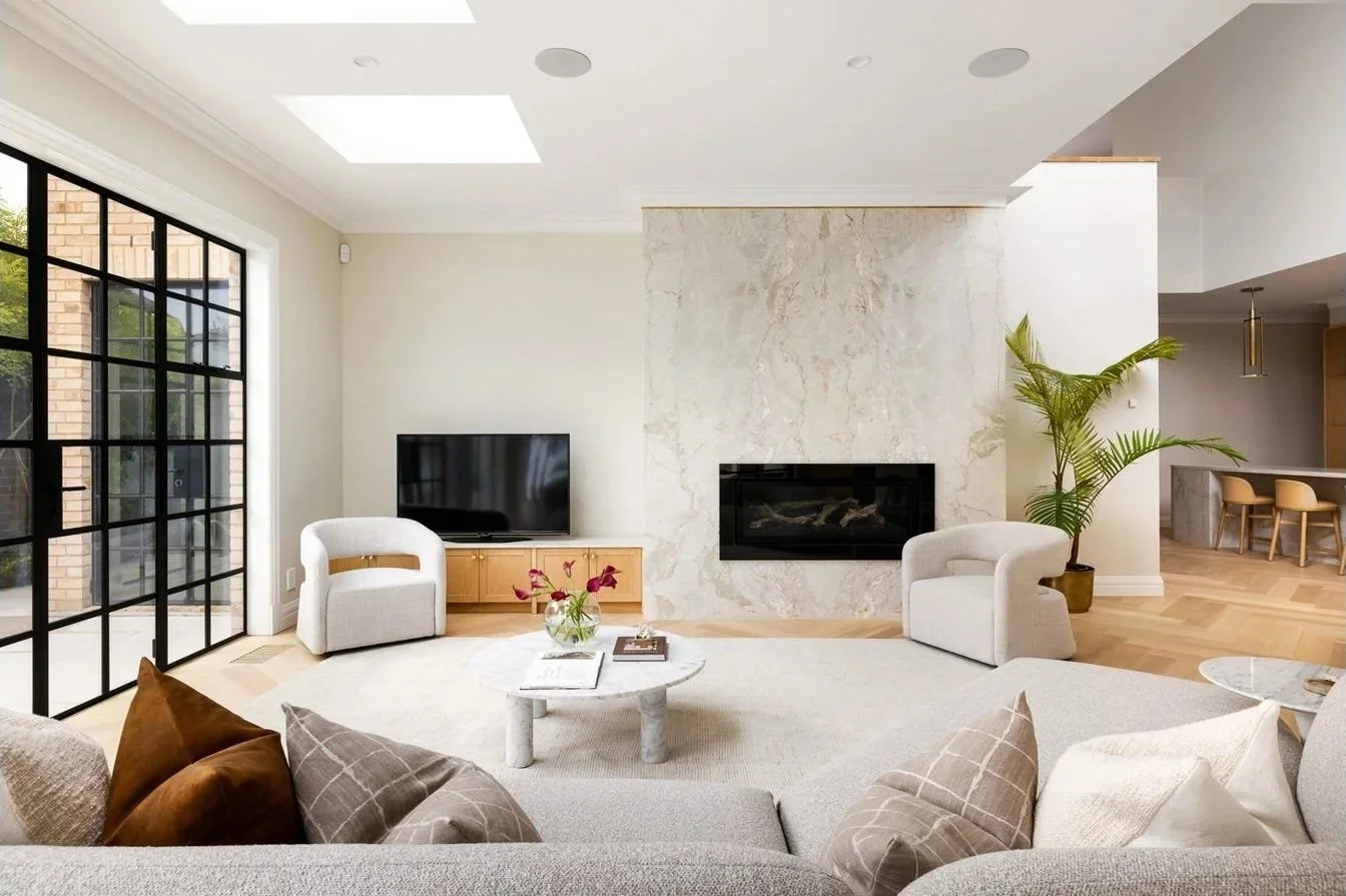 Modern living room with white and beige decor, black-framed glass sliding doors, marble fireplace, white armchairs, a round coffee table with flowers, a TV, and a potted plant.