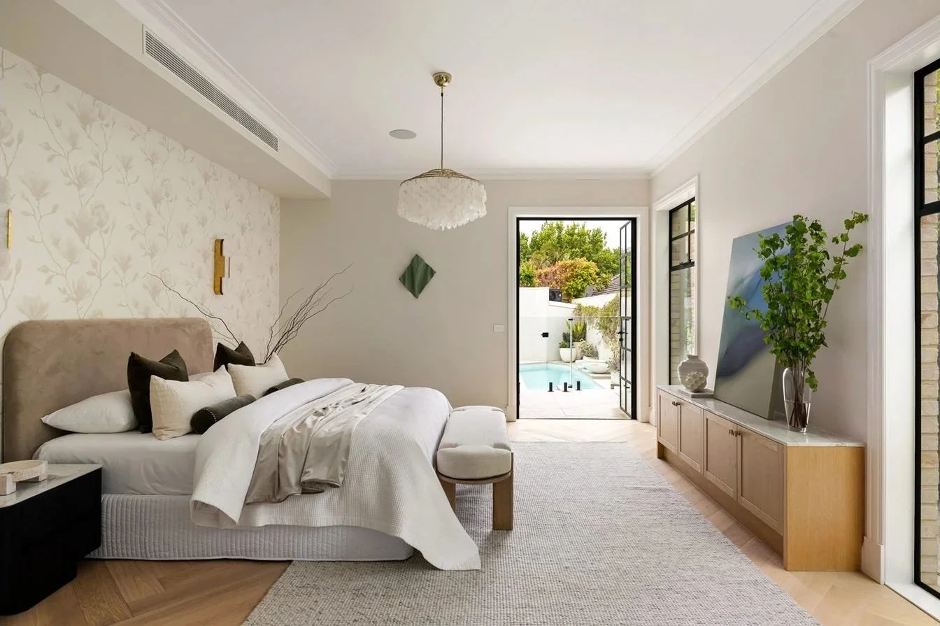 Bright bedroom with large open door to outdoor pool scene, neutral decor, bed with beige headboard, white and beige bedding, various pillows, light wood furniture, artwork, mirror, and potted plant.