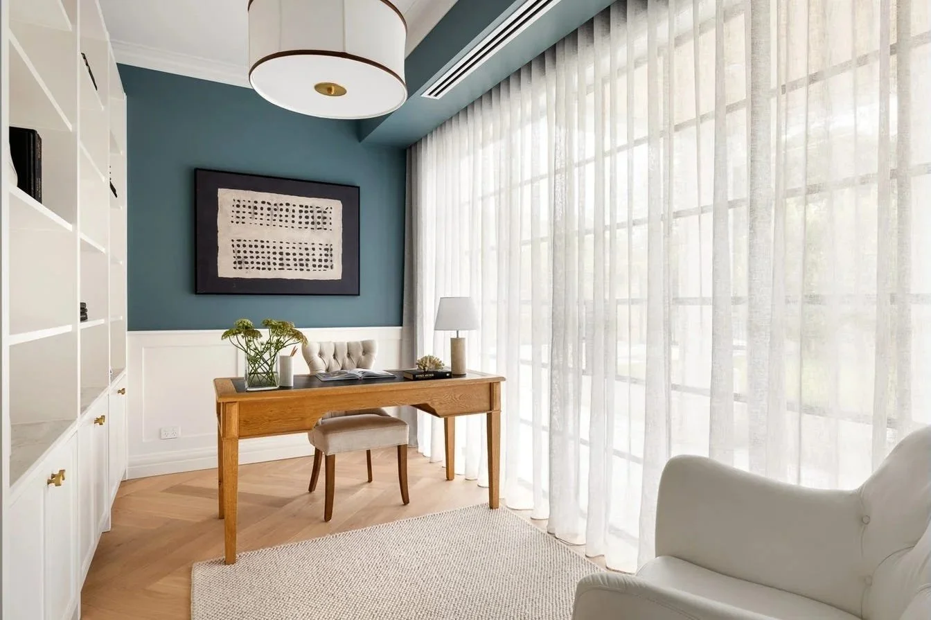 A cozy home office with a wooden desk, a beige upholstered chair, a white armchair, white sheer curtains covering large windows, and a modern pendant light fixture. There's a dark framed artwork on a blue wall, a white bookshelf, and a plant on the d