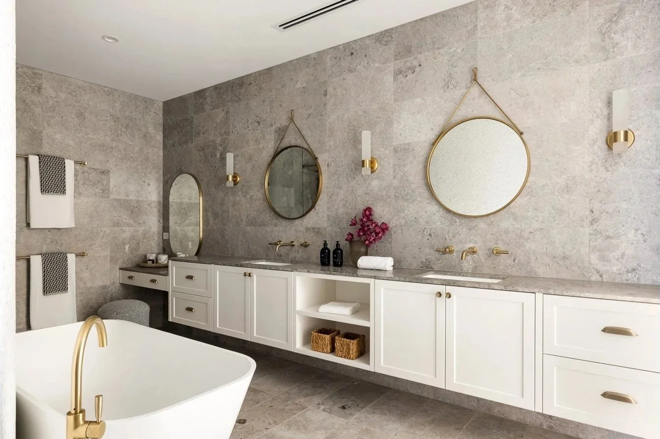 Modern bathroom with gray stone tiles on walls and floor, white vanity with gold handles, oval and round mirrors, gold light fixtures, a bathtub with a gold faucet, towels, and decorative baskets.