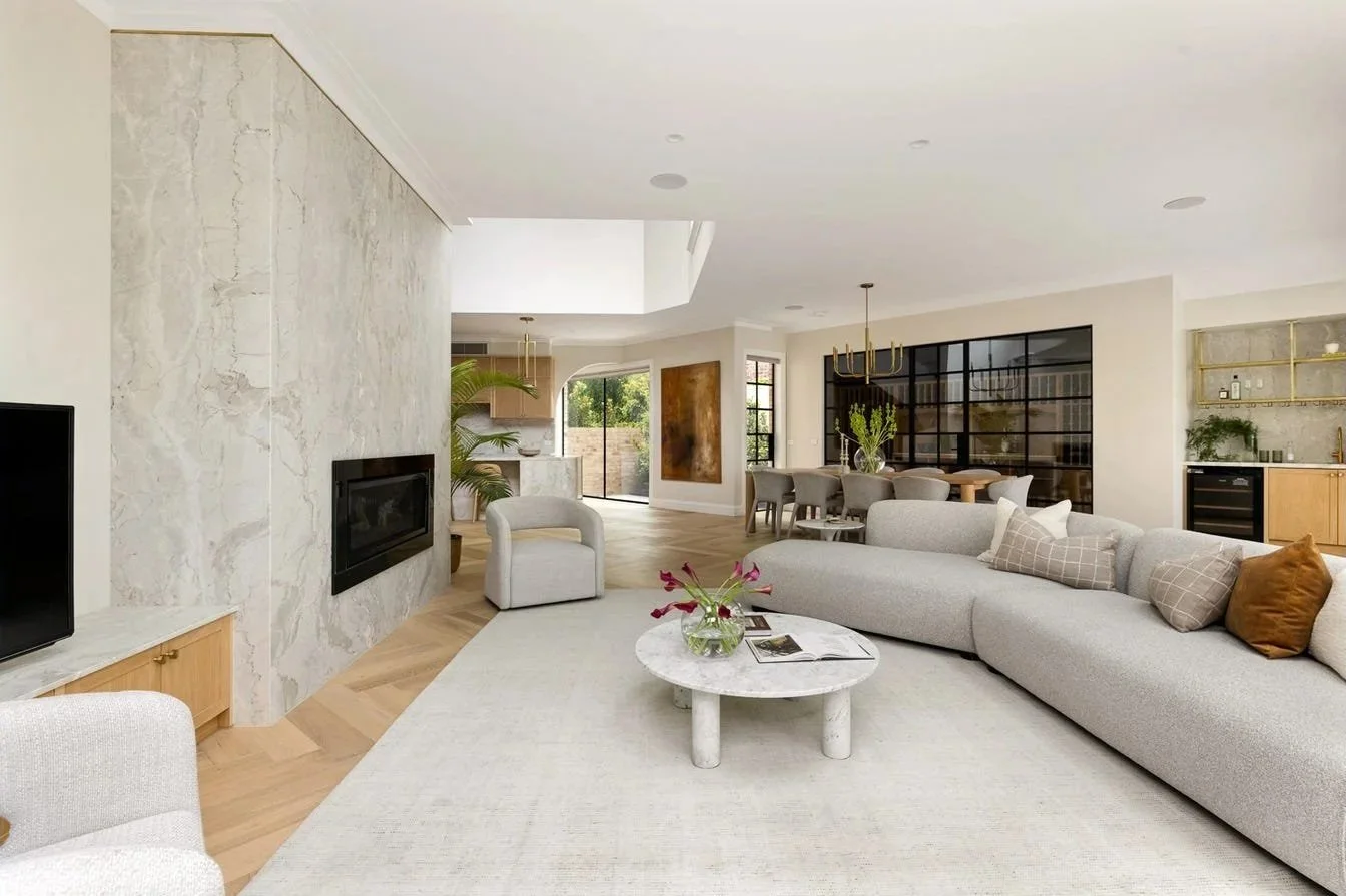 Spacious modern living room with light-colored sectional sofa, marble coffee table with flowers, flat-screen TV, fireplace, dining area with round table and chairs, and a kitchen in the background.