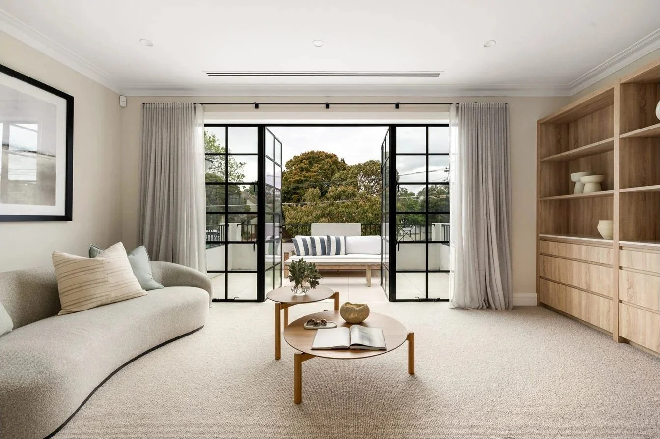 Bright living room with open balcony doors, beige sofa, light carpet, wooden coffee tables, and built-in wooden shelving unit.