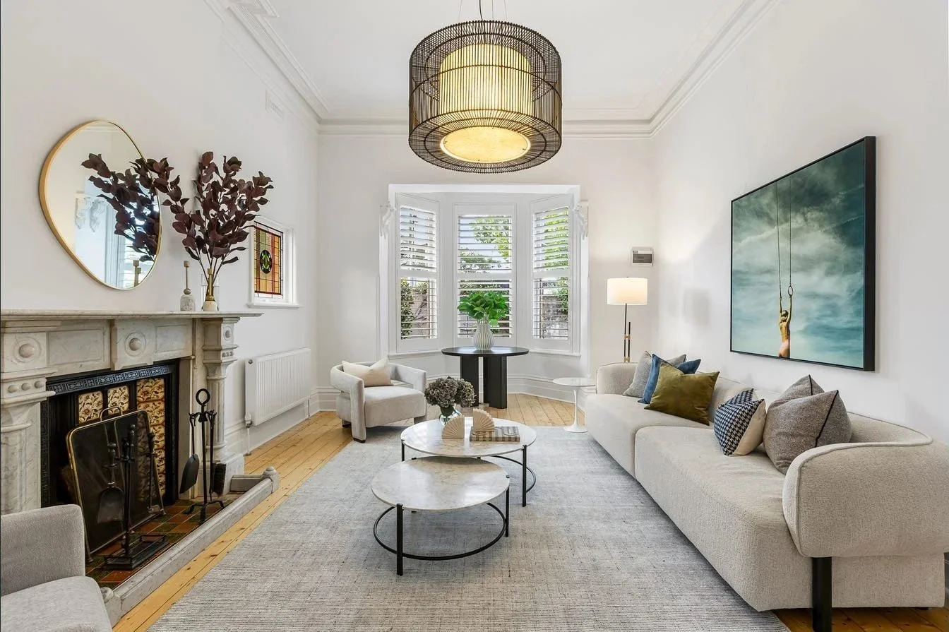 Bright living room with a fireplace, large window with shutters, modern beige sofa with cushions, round marble coffee tables, plant on window ledge, floor lamp, wall art depicting a woman on swing, and decorative items on mantel