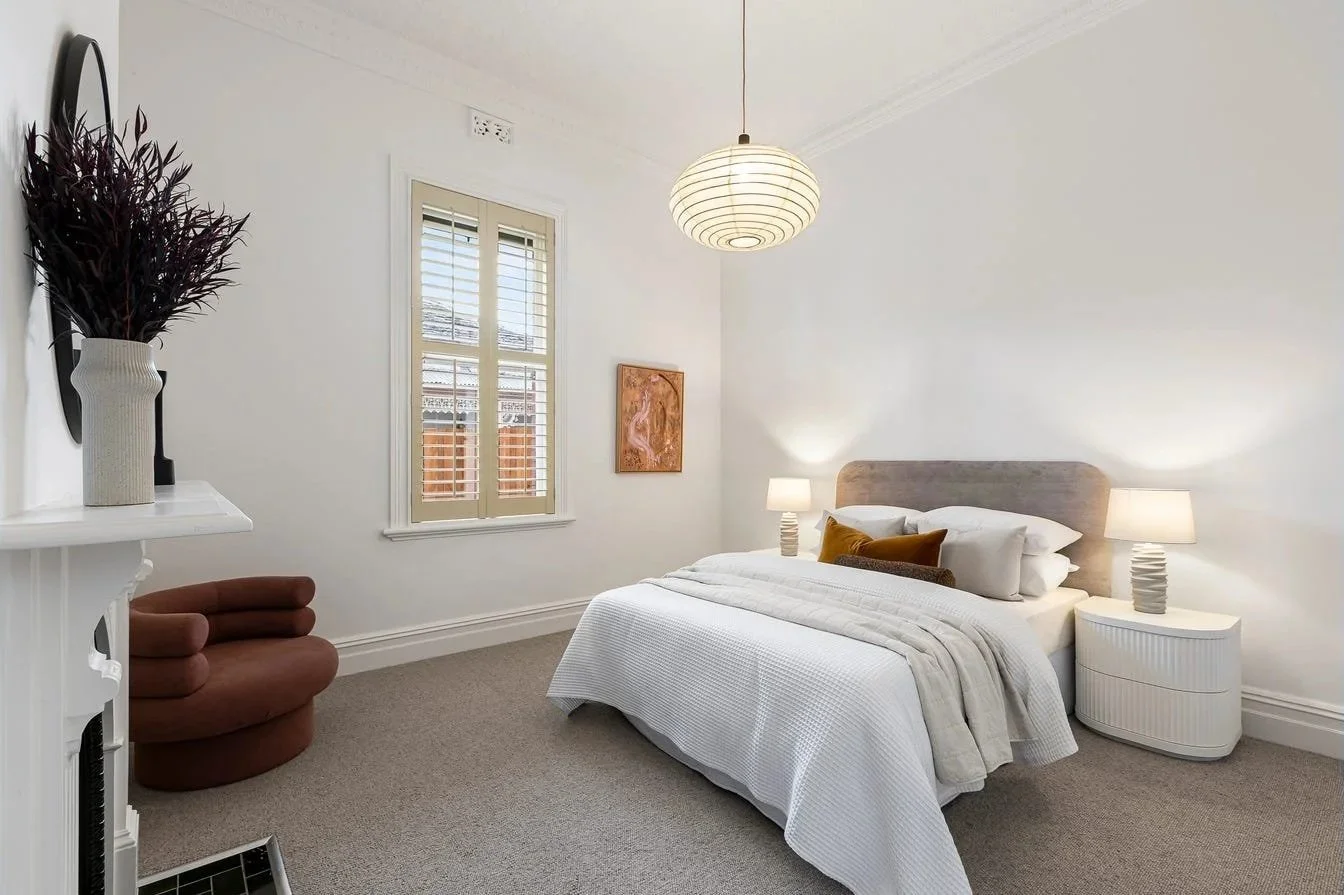 A bedroom with a white bed, beige headboard, two white nightstands with lamps, a window with blinds, a pink abstract painting, a brown upholstered chair, and a white fireplace mantel with a vase of dark purple flowers.