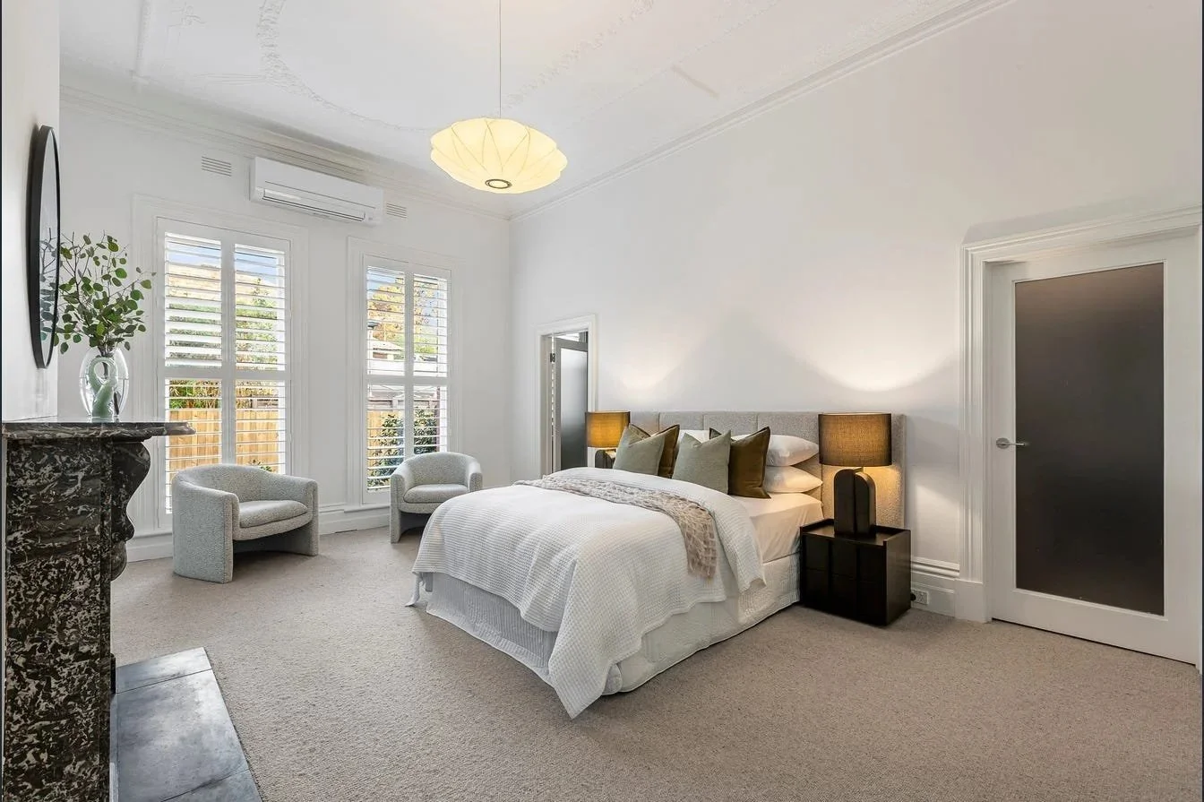A bright, modern bedroom with white walls, a large bed with white bedding and multiple throw pillows, two bedside lamps, a dark cabinet, a marble fireplace, two armchairs near windows with plantation shutters, and a ceiling light fixture.